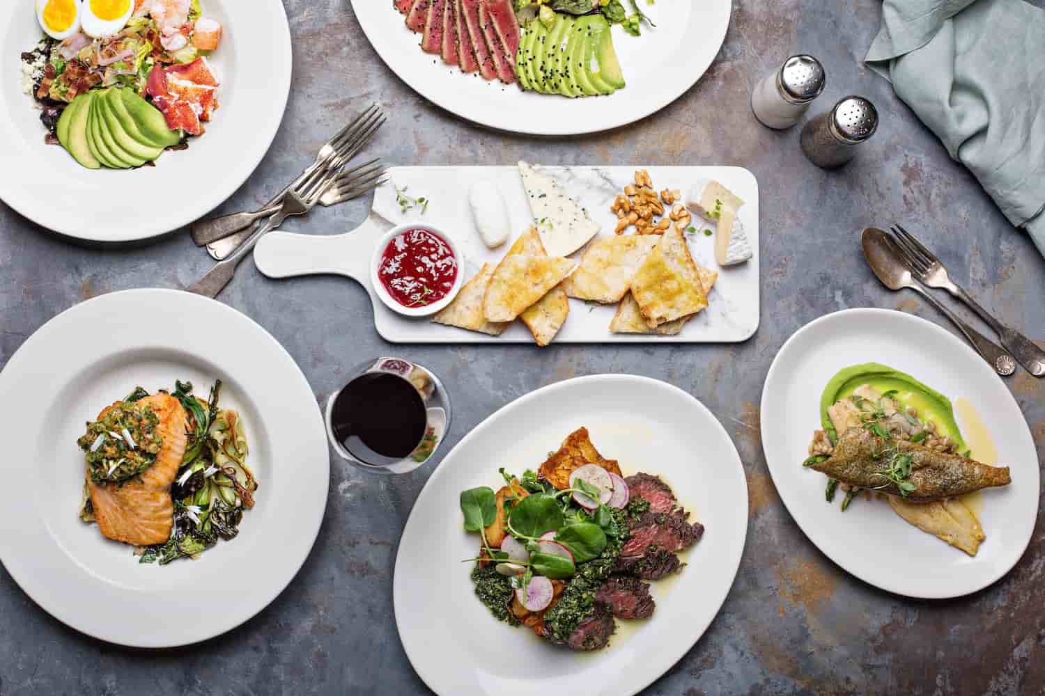 Uma mesa com diversos pratos de comida em cima, com alguns talheres e temperos em volta.